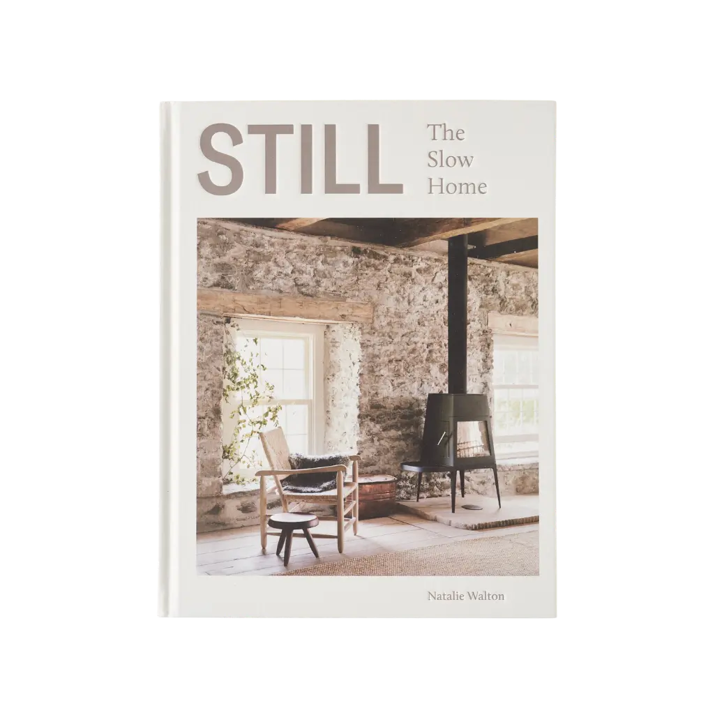 Still: The Slow Home by Natalie Walton, a book about creating a peaceful, grounded sanctuary. Book cover showing a rustic interior with thick stone walls, exposed timber beams, and a wood-burning stove, evoking a sense of warmth, history, and quietude.