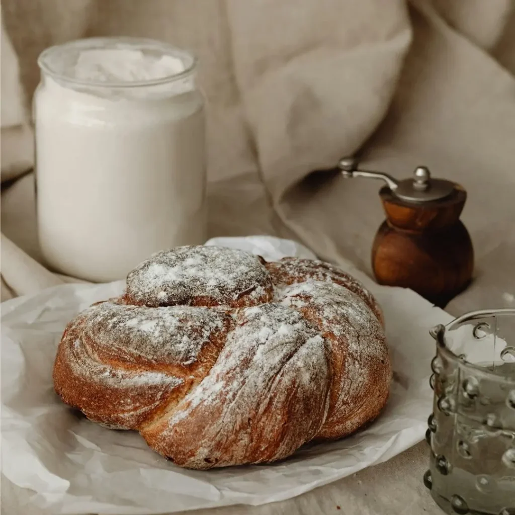 A loaf of braided artisan bread dusted with flour on parchment paper, next to a jar of flour and a wooden pepper mill.