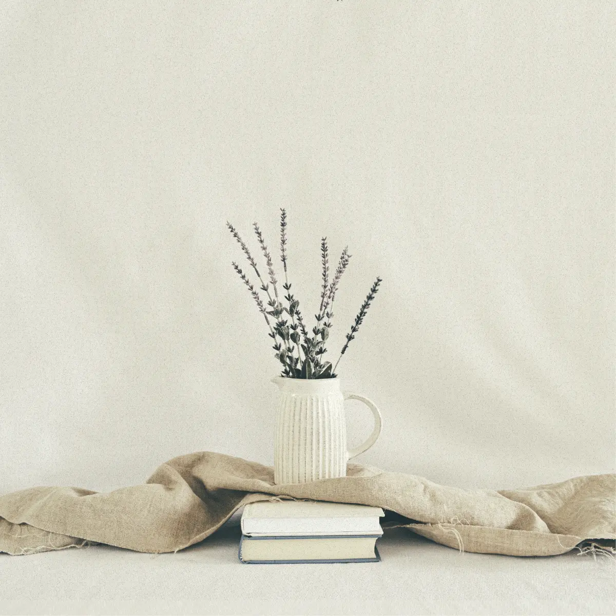 A white ceramic jug with vertical ridges sits on a small stack of books. The jug holds a few stems of dried lavender. A piece of natural linen fabric is draped loosely behind and around the books. The entire scene is set against a soft, neutral background in a minimalist, vintage-inspired style.
